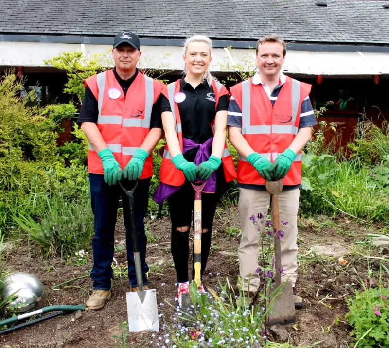 Team of people standing in middle of garden patch, wearing high-vis vests and holding spades.