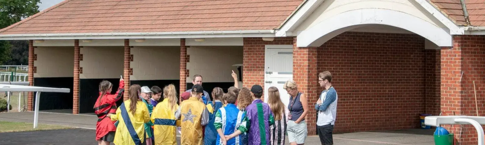 Group of children wearing jockey silks standing in front of stables.